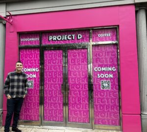Man stood in front of a pink shop with ProjectD Doughnut logo on